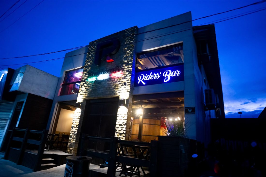 Fachada da Pandorato Pizzaria e do Riders Bar à noite, com o letreiro azul do bar