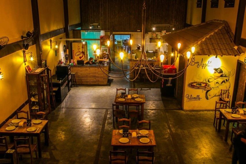 Salão da pizzaria visto de cima, com mesas de madeira e lustres de luz quente